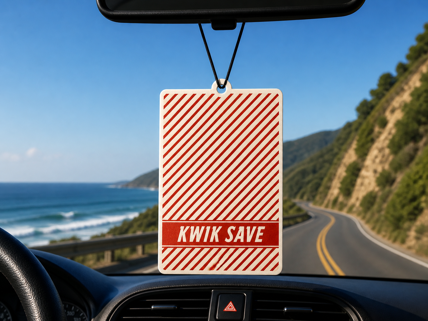 Kwik Save air freshener hanging from a car's rearview mirror with a scenic road and ocean view outside.