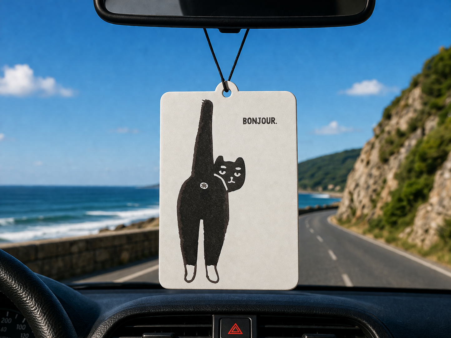 Car air freshener with a cat illustration hanging from a car's rearview mirror, with a scenic road and ocean view outside.