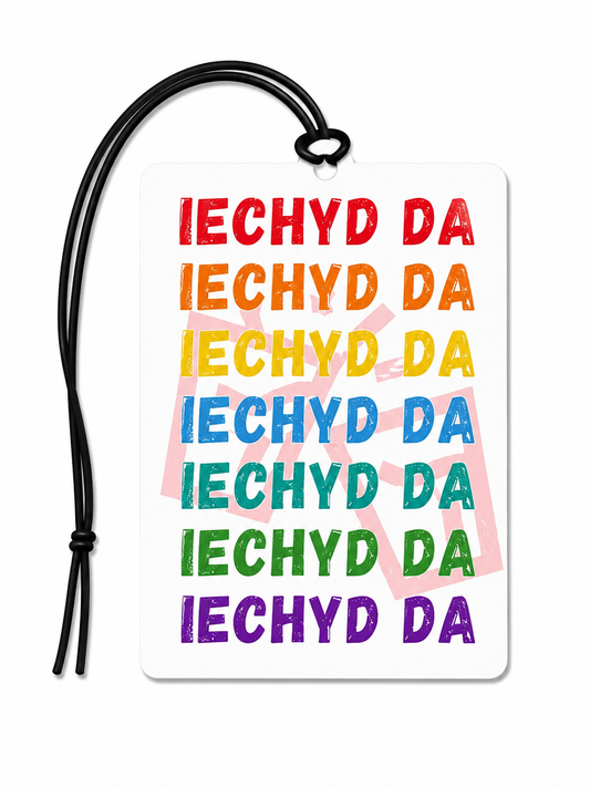 A rectangular white car air freshener with rounded corners, featuring the phrase 'IECHYD DA' repeated in stacked rows of colorful text (red, orange, yellow, blue, teal, green, purple) on a white background. A black hanging cord is attached at the top, and a faint pink 'DO NOT DISTURB' watermark is visible behind the text.