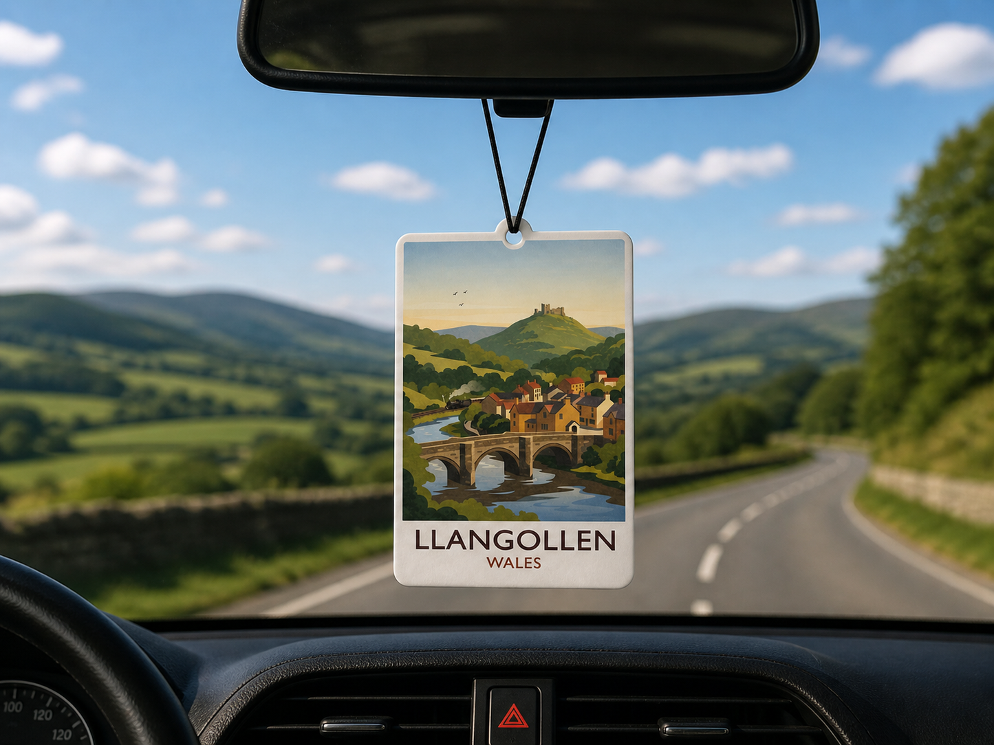 Car air freshener with Llangollen, Wales design hanging from a car's rearview mirror with a scenic view of the town.