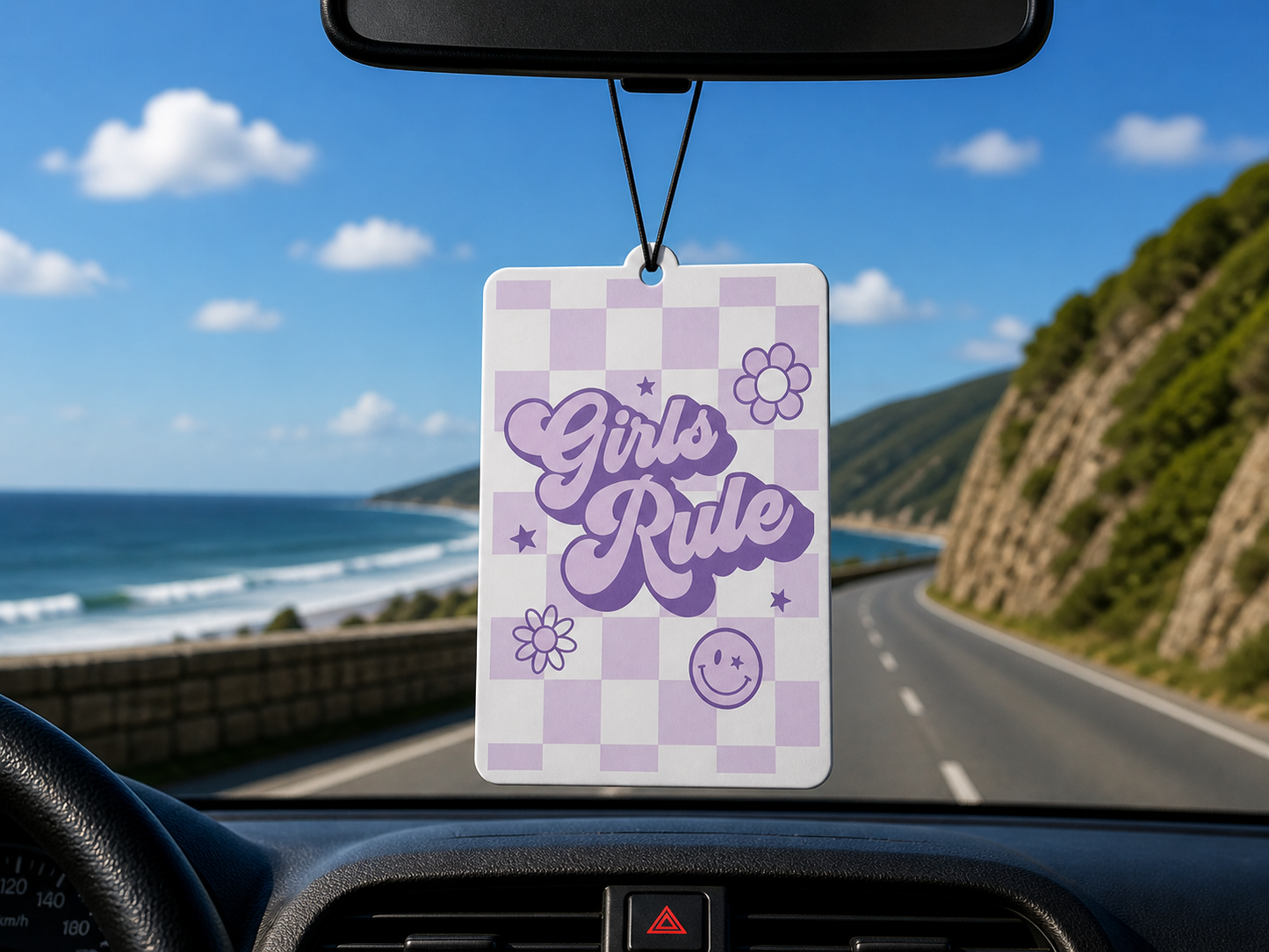 Car air freshener with 'Girls Rule' text hanging from a car's rearview mirror, with a scenic road and ocean view outside.
