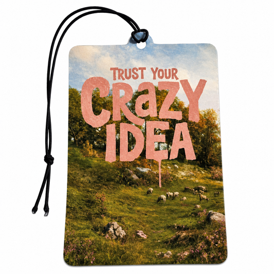 Trust Your Crazy Idea motivational car air freshener featuring pink lettering over a scenic countryside landscape with sheep and trees.