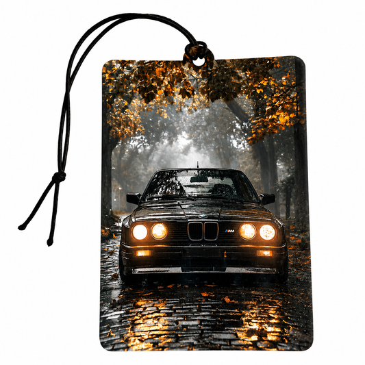 A rectangular car air freshener or luggage tag with a black hanging cord, printed with a front-facing black BMW M4 in a misty autumn forest with glowing headlights and golden leaves.