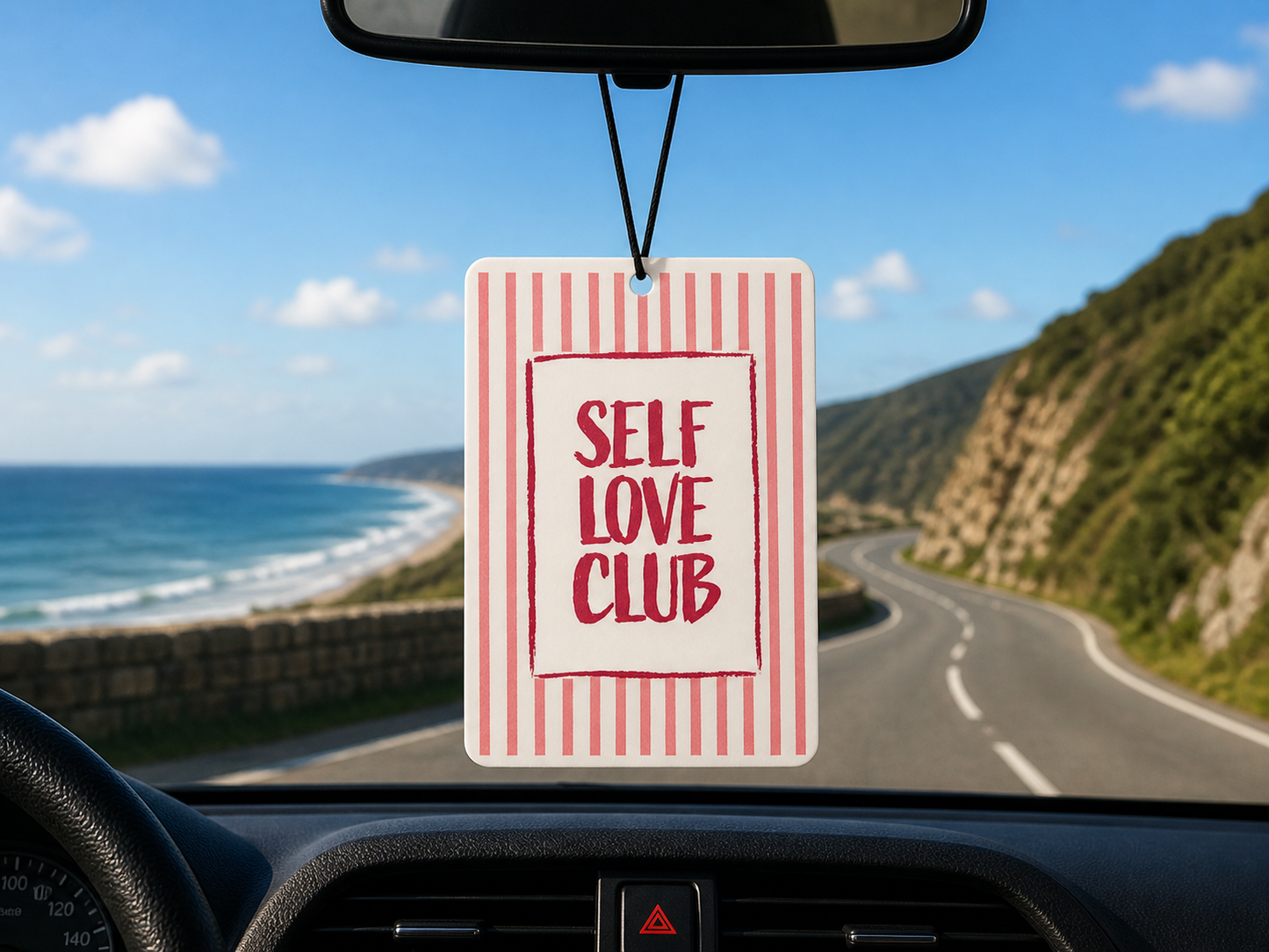 Car air freshener with 'Self Love Club' text hanging from a car's rearview mirror, with a scenic road and ocean view outside.