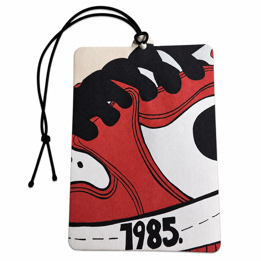 A rectangular car air freshener tag featuring a graphic print of a red, black, and white sneaker with the year '1985.' printed on it.